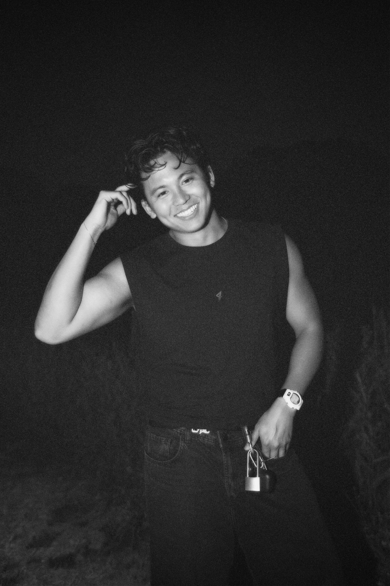 Black-and-white photo of Alastair smiling broadly in the dark, posing with a hand to his head in a tank top, watch, and carabiner clip.