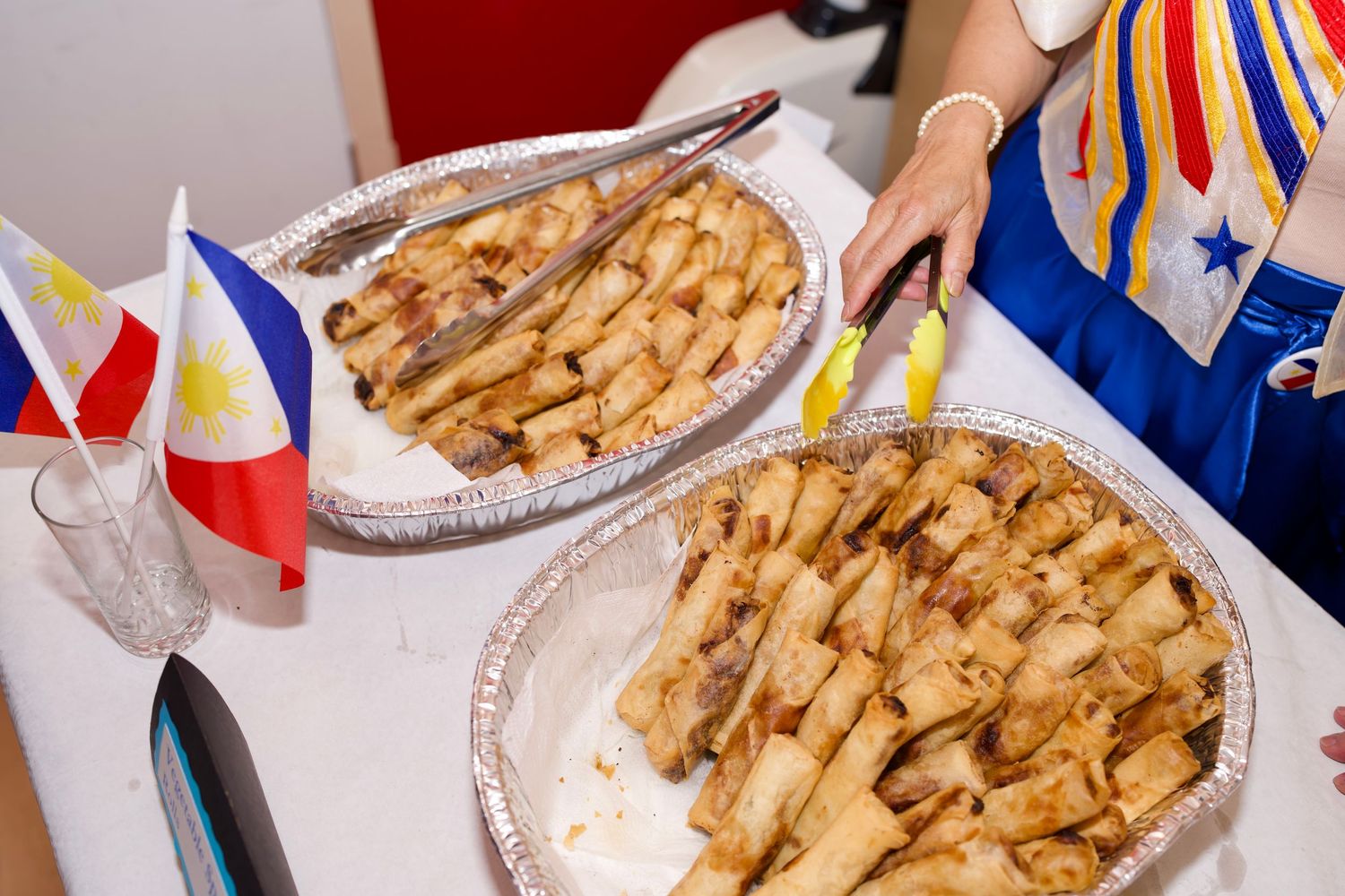 Filipino community gathering around an indoor dining table enjoying shared meals and holding handheld flags with spring rolls.