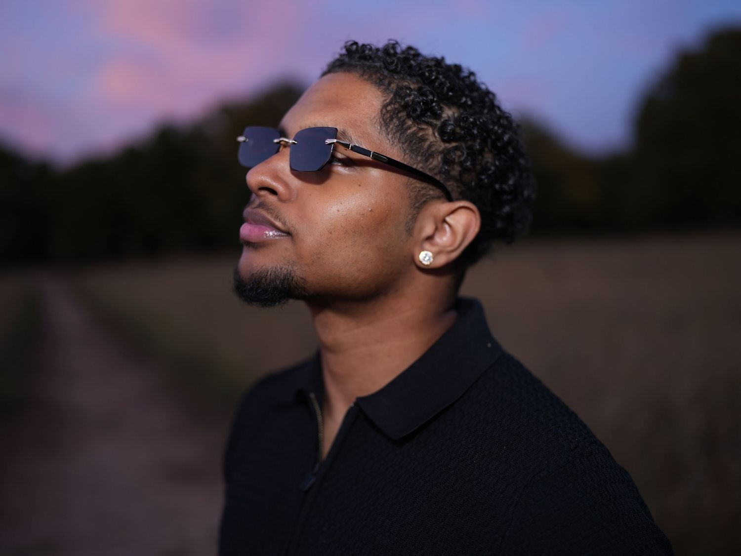 Profile portrait of a man in sunglasses looking upward against a dusk sky.