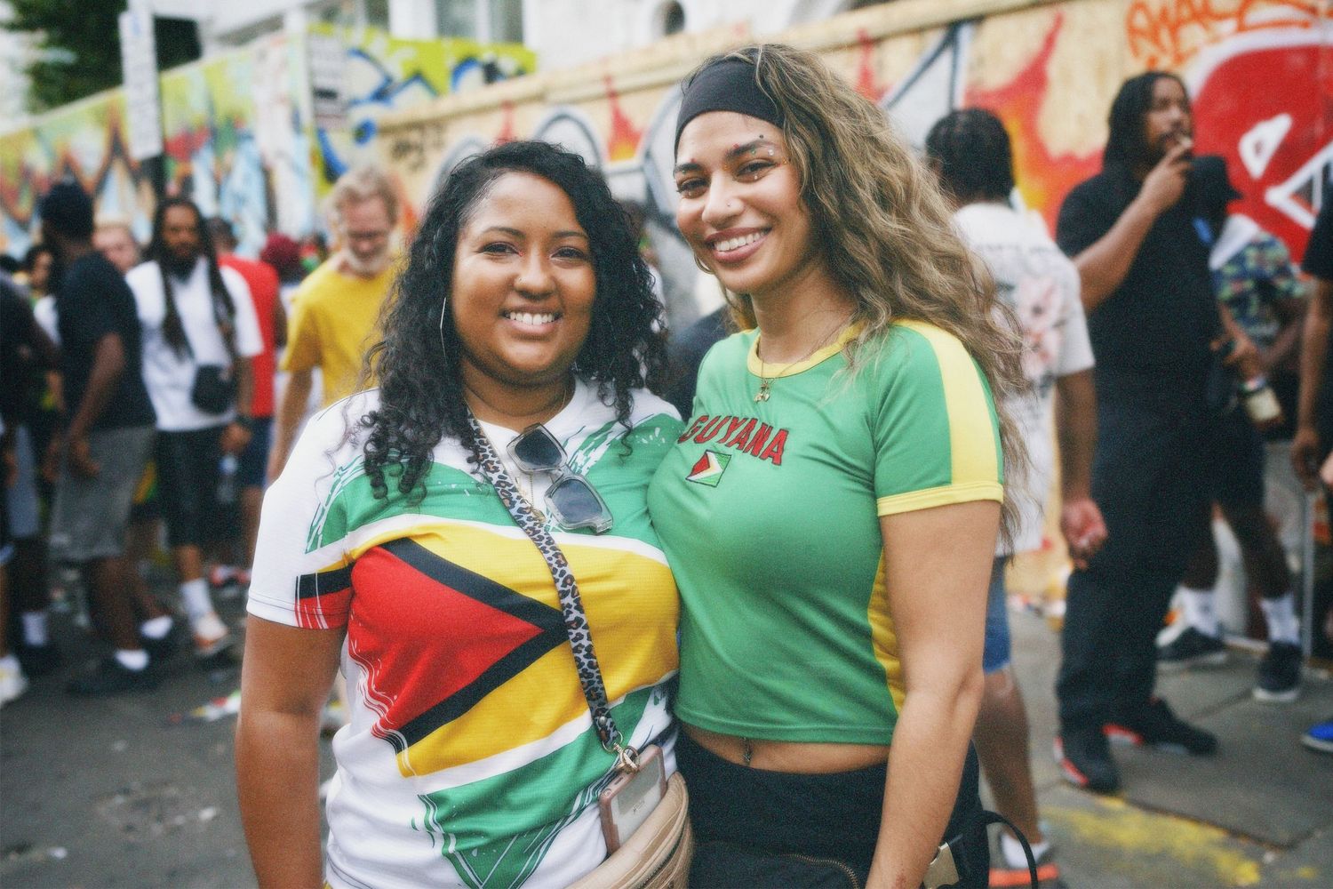 Two women smiling in vibrant Guyana flag t-shirts on the road at Notting Hill Carnival.