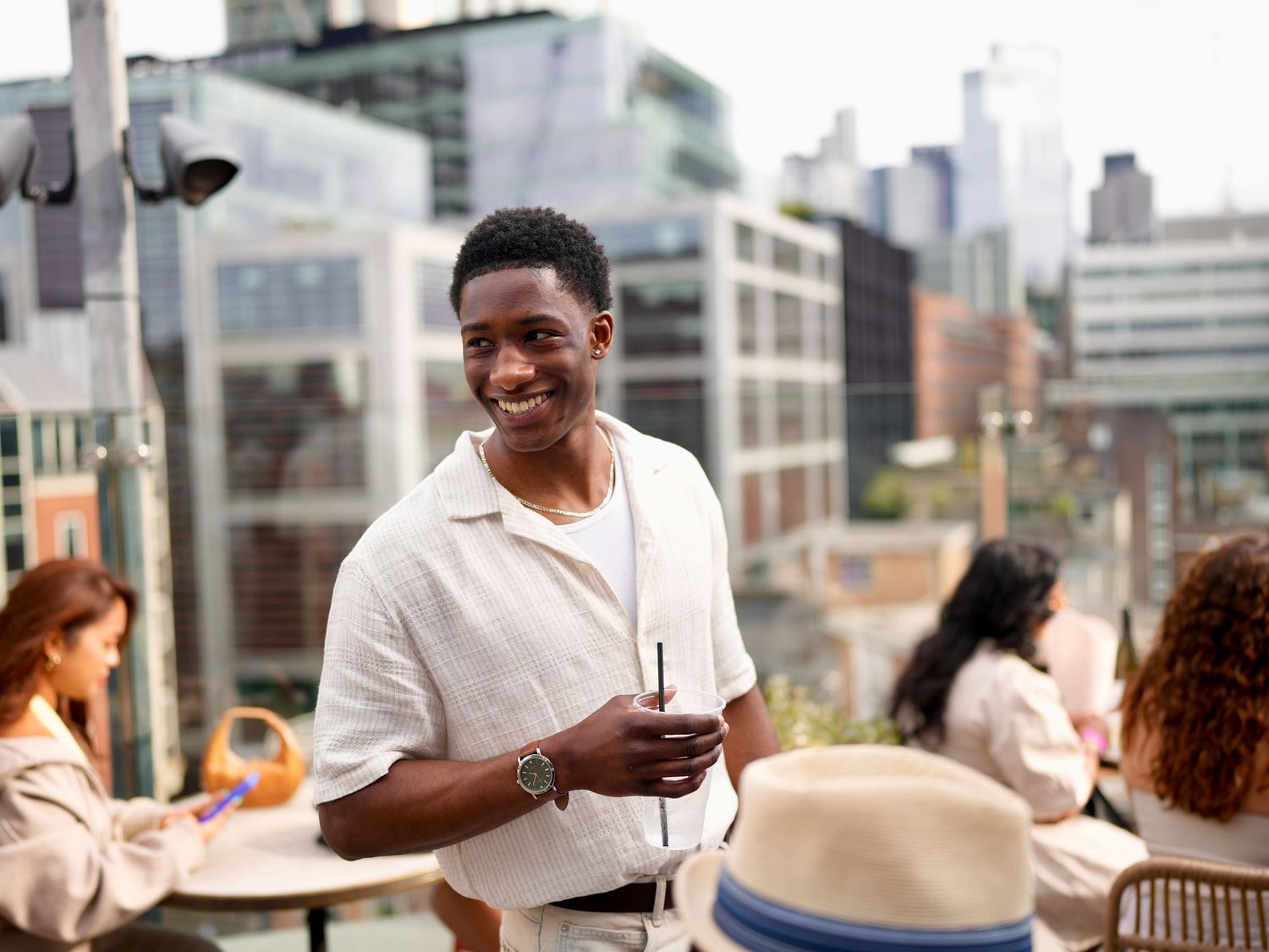 A smiling man holding a drink at a bustling outdoor rooftop venue with city views.