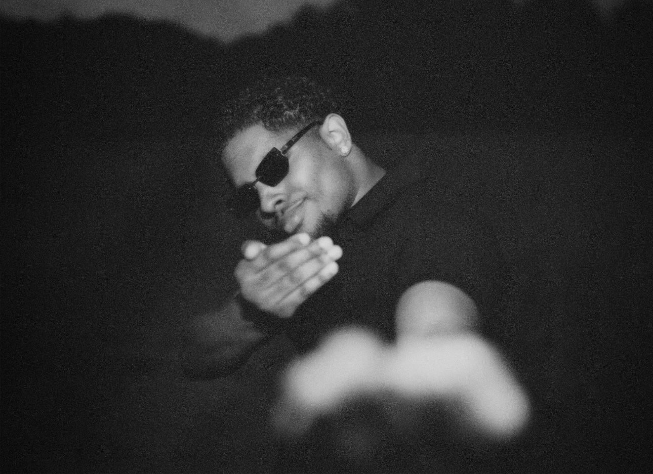 Gritty black-and-white portrait of a man in sunglasses reaching out towards the camera with out-of-focus hands in the foreground.