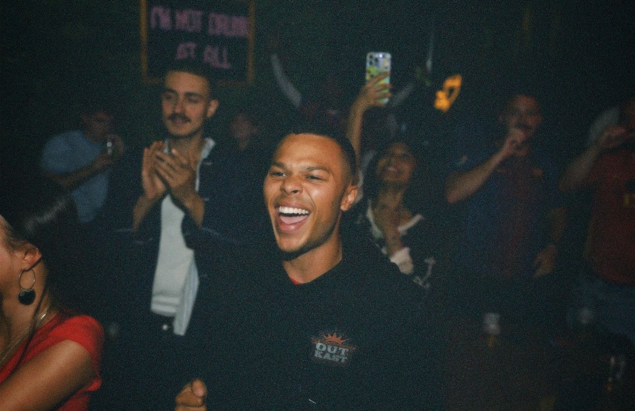 A man in a black hoodie cheering enthusiastically during a live sports screening event by ARIETA Studios.