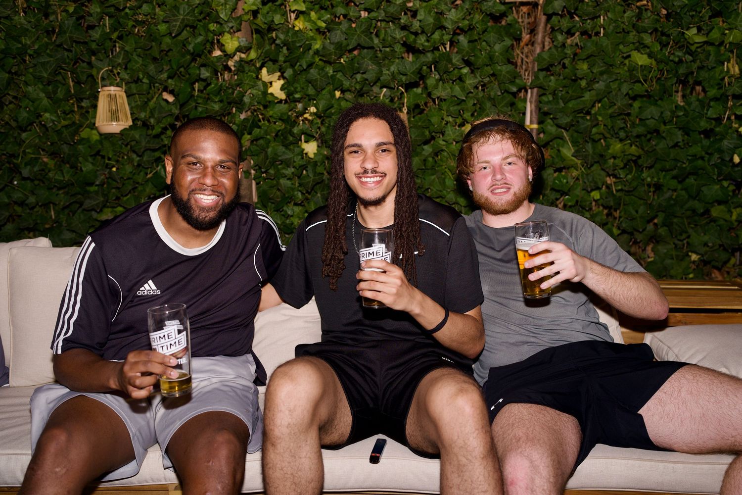 Three men smiling and laughing on a couch while holding drinks at an outdoor evening social. Photo by ARIETA Studios.