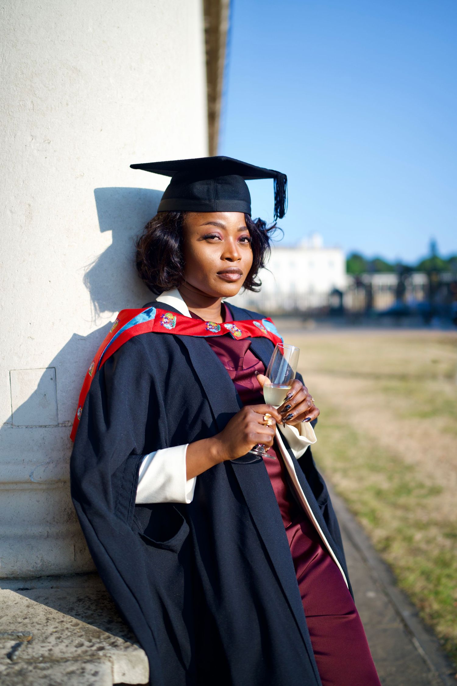 Graduate in academic cap and gown leaning on stone pillar at Greenwich graduation shoot by ARIETA Studios.
