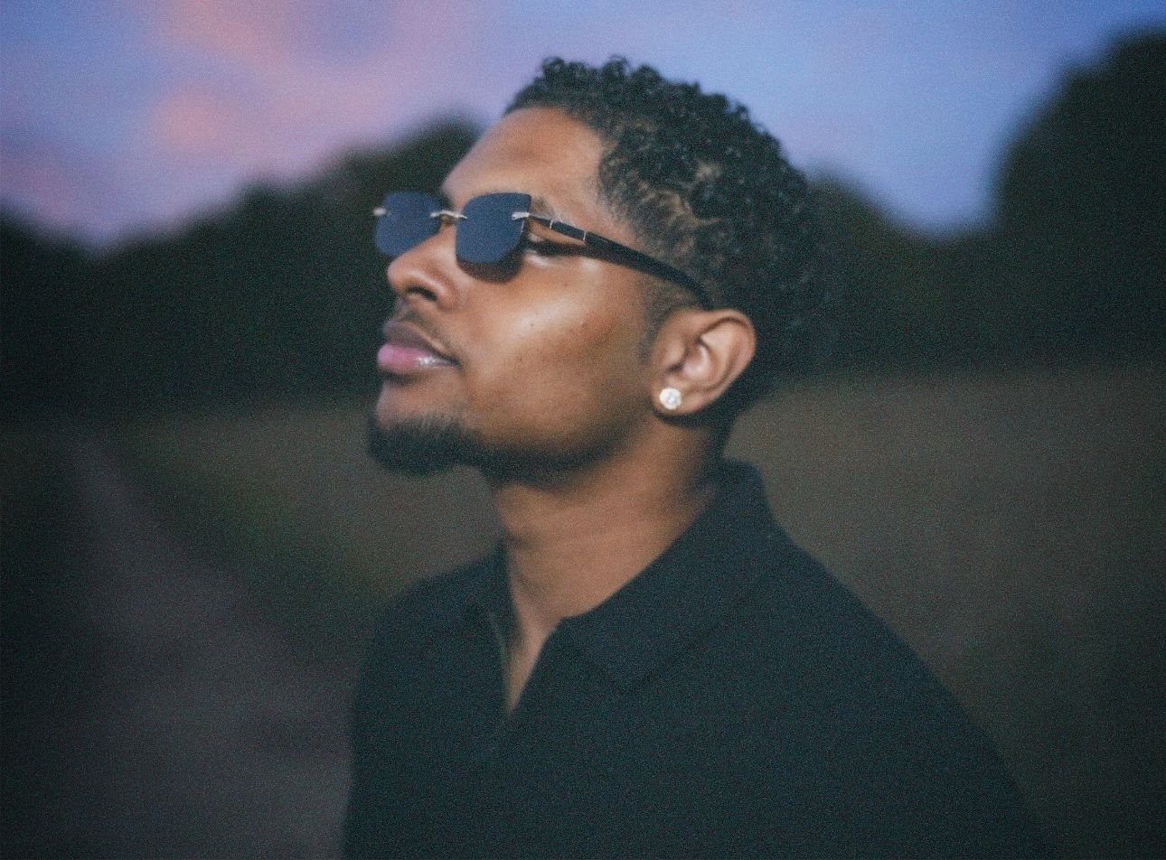 Moody, close-up side profile portrait of a man wearing rimless sunglasses and a diamond stud earring, illuminated by soft ambient low light.