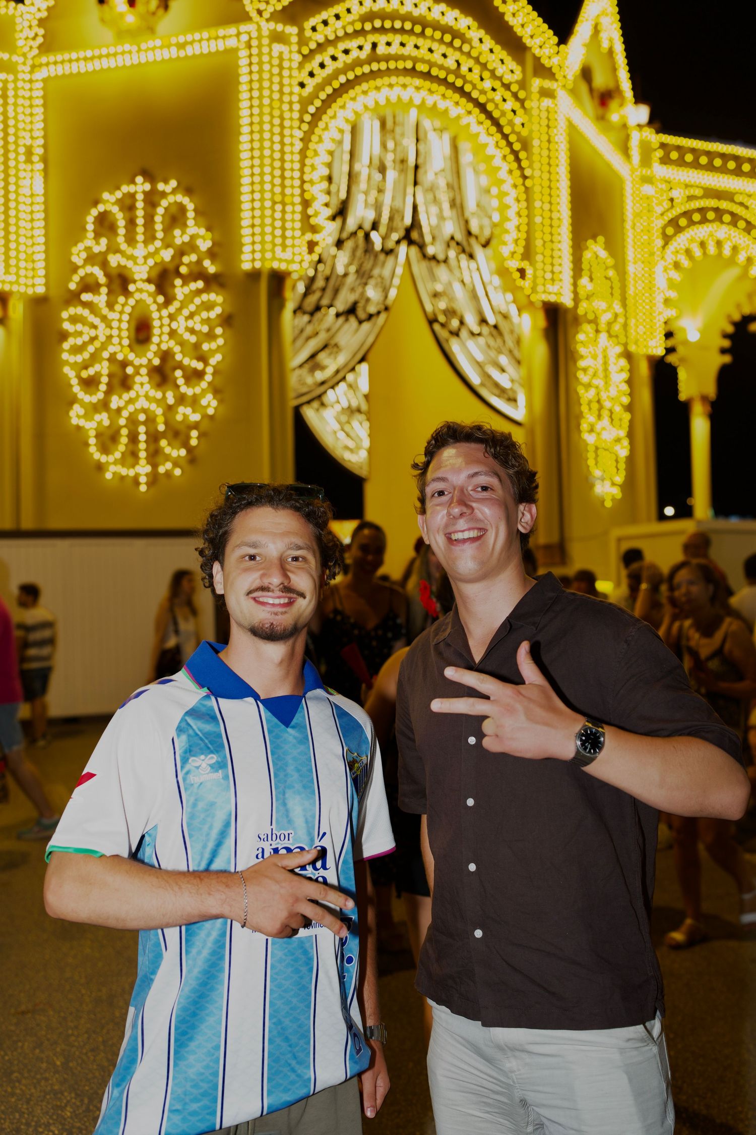 Two guys smiling under glowing festival lights at Feria de Málaga / Dos chicos sonriendo bajo las luces de la Feria de Málaga