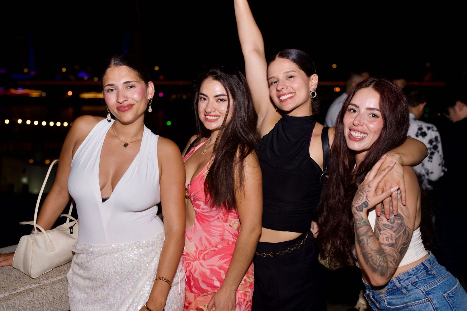 Four women dressed in chic party outfits smiling and hugging on the outdoor deck against the night sky.