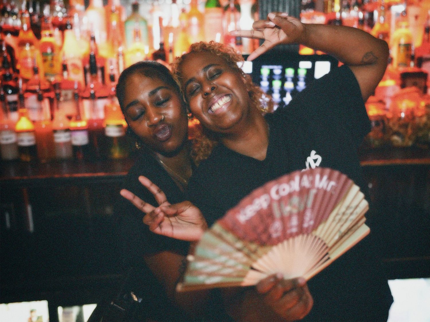 Two women in black branded crop tops posing in front of lit bar shelves by ARIETA Studios