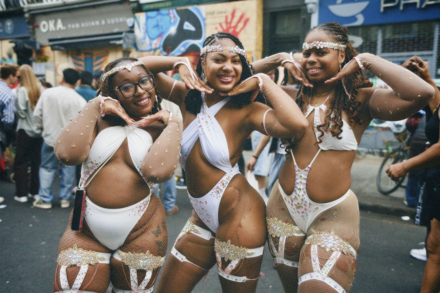 Three women striking signature hand poses in white festival outfits at Notting Hill Carnival.