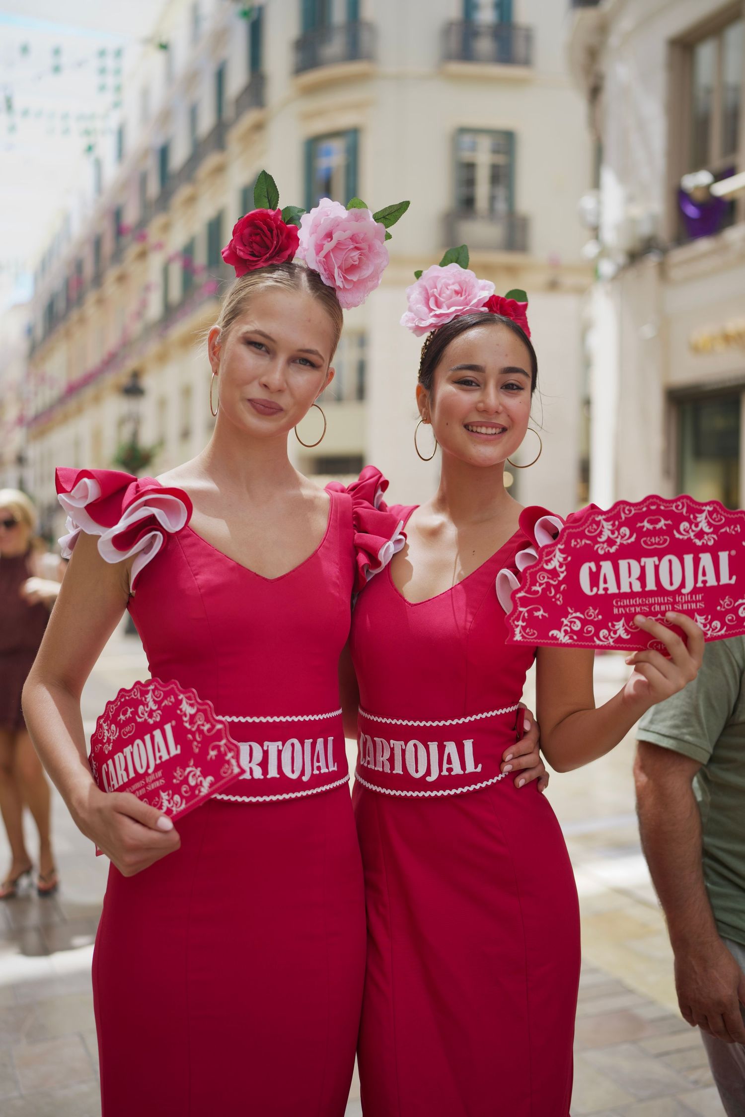 Two women in matching pink dresses with floral headpieces holding Cartojal fans on the sunny streets of Feria de Málaga.