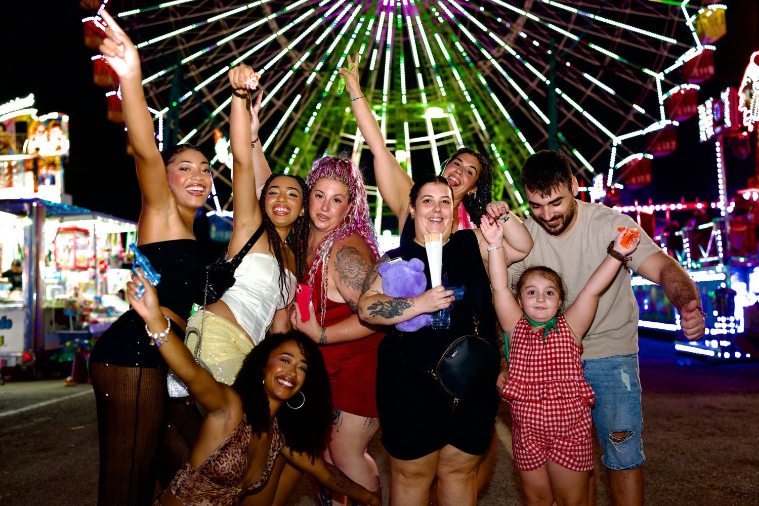 Group of friends cheering at night in front of an illuminated carnival ride at Feria de Málaga.