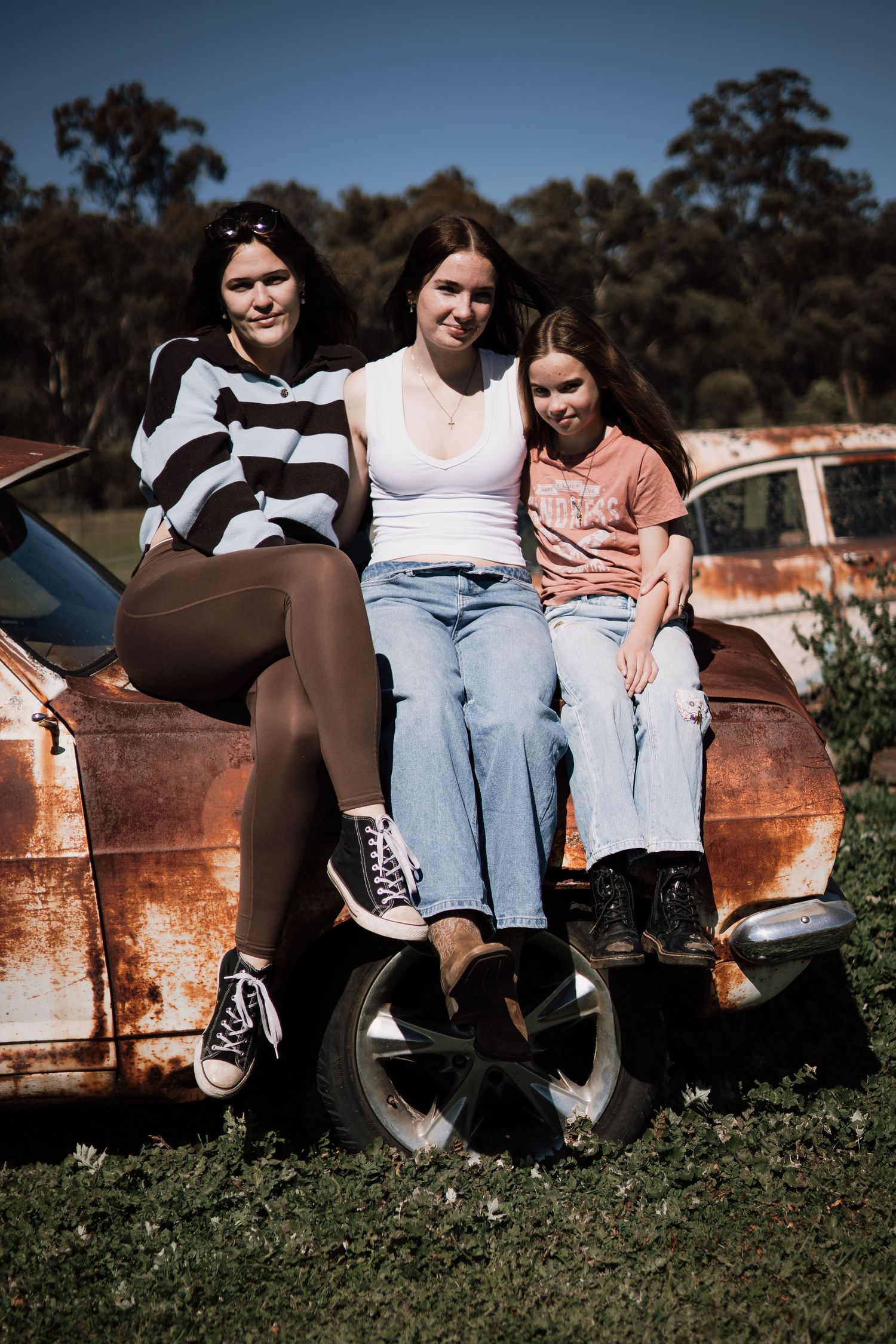 Three young women posing together on a rusty vintage car in a rural outdoor setting on a sunny day.