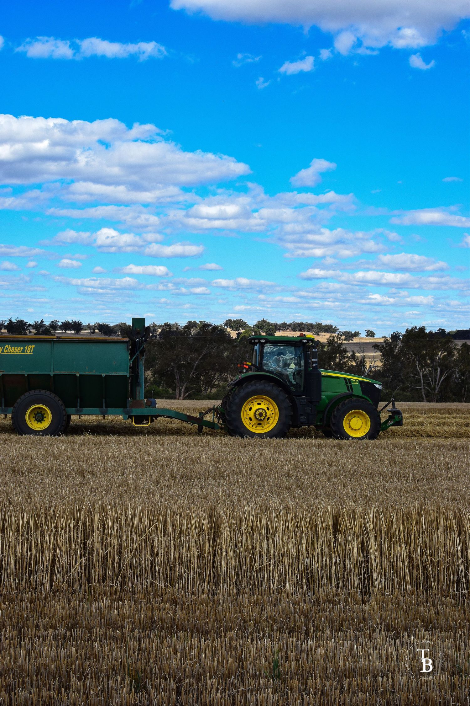 Green John Deere tractor pulling grain cart across harvested wheat field under blue sky.