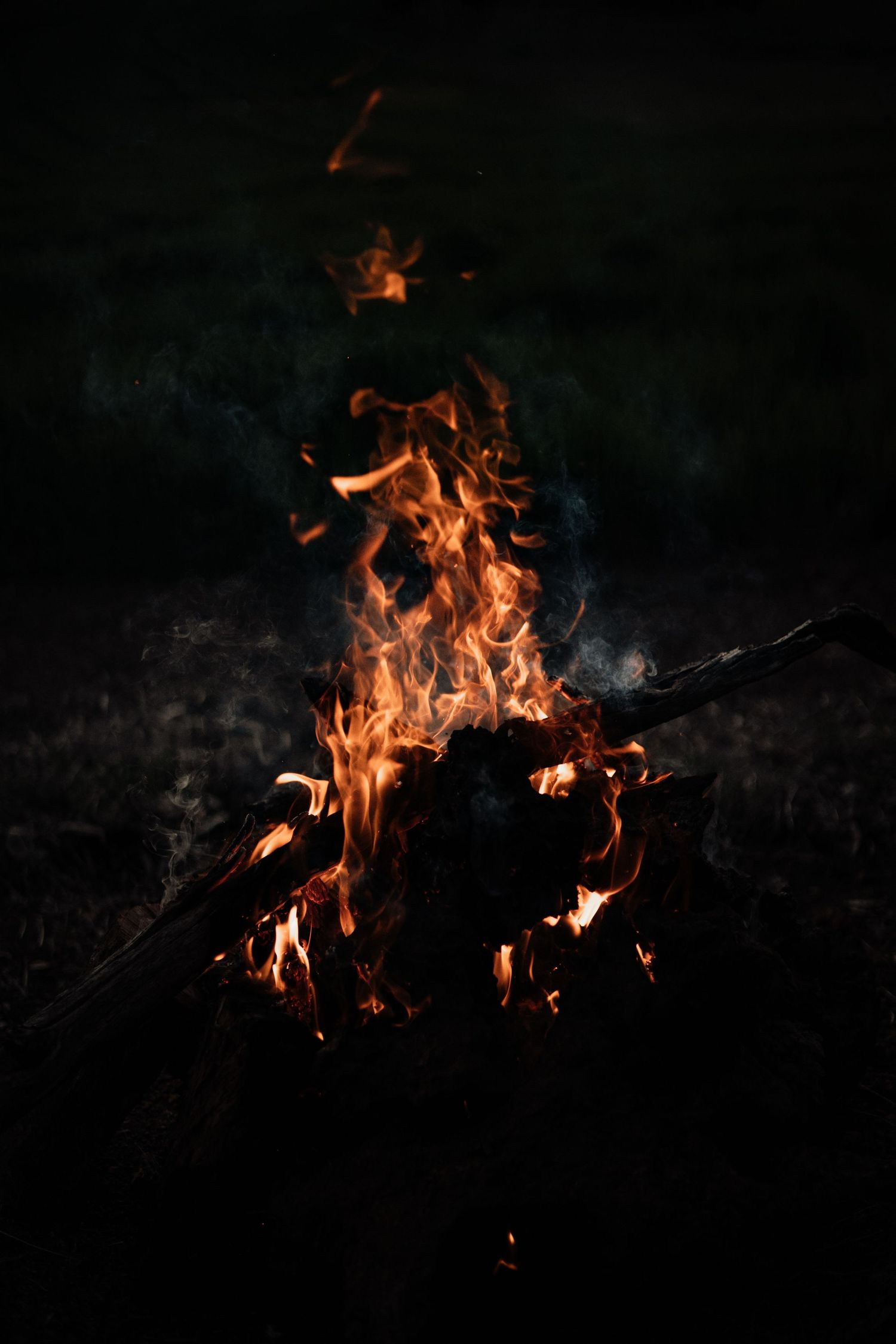 A glowing campfire burns brightly against a dark night sky, with orange flames and smoke rising from wooden logs.