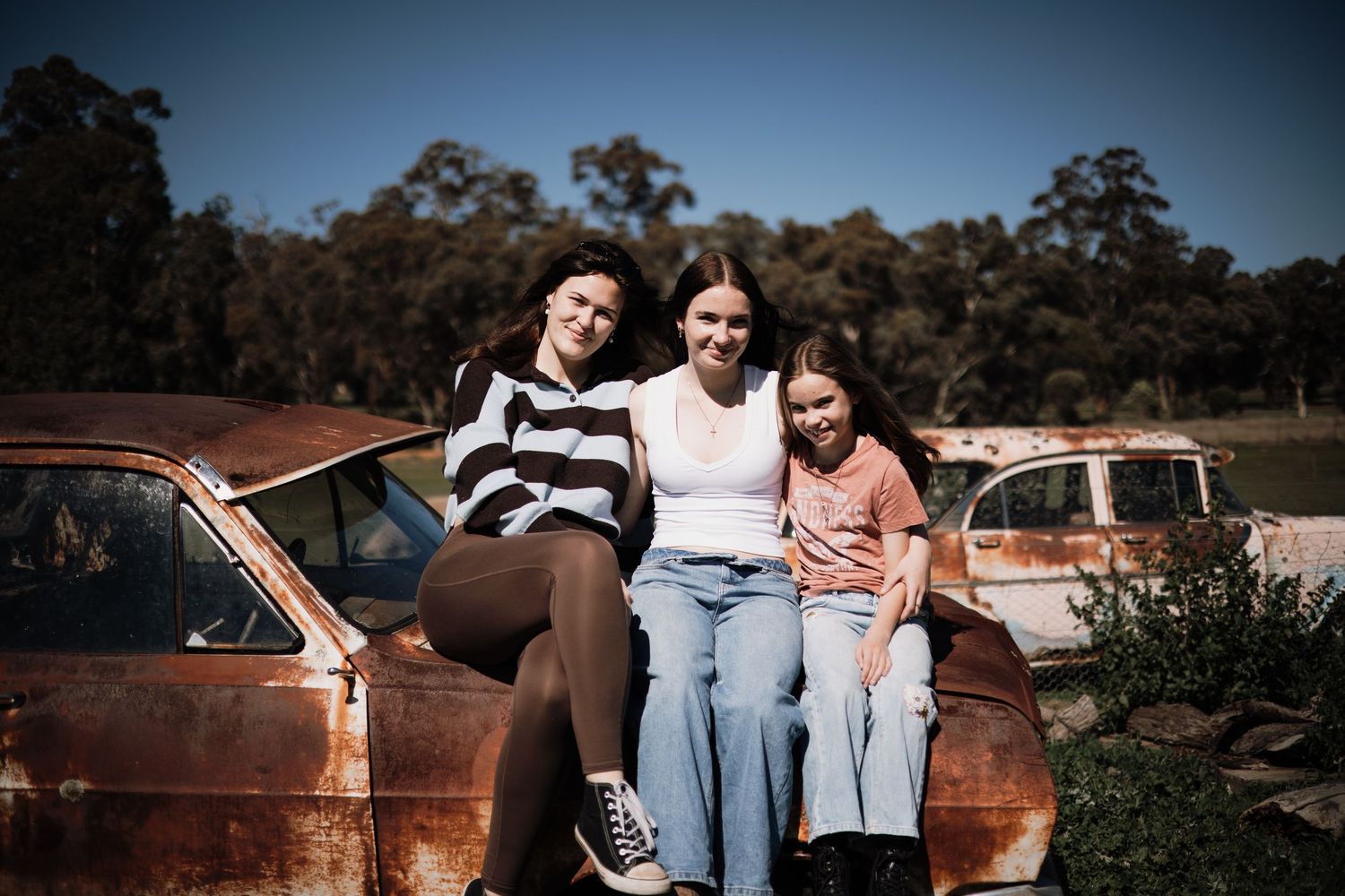 Three young women posing together on a rusty vintage car in a rural outdoor setting on a sunny day.