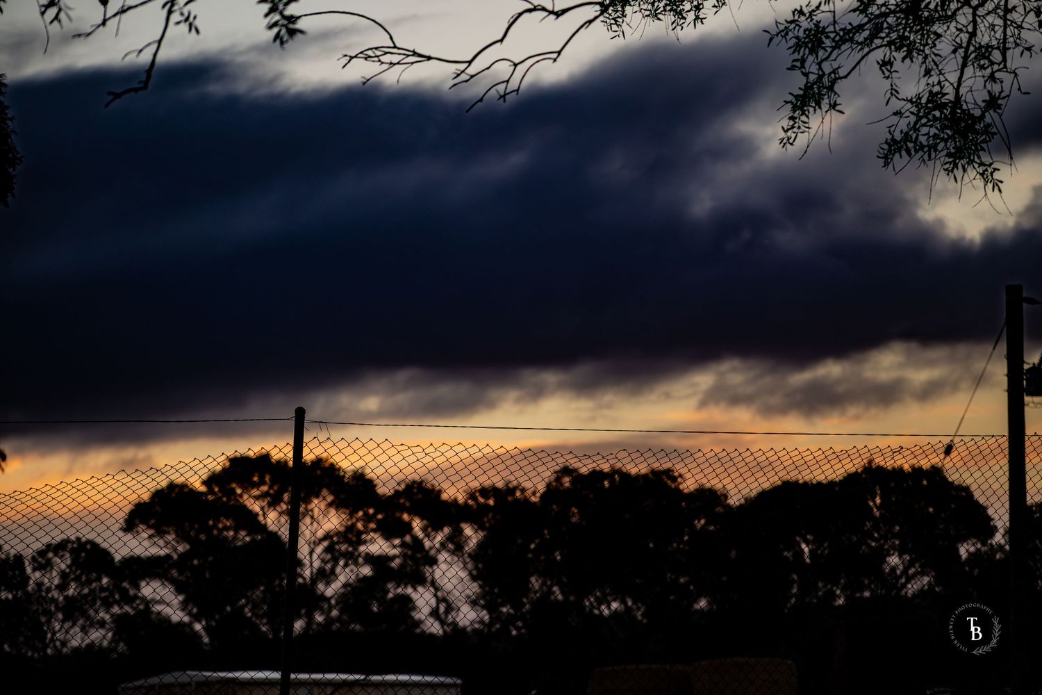 Dramatic dark storm clouds silhouette trees and a wire fence at dusk with orange sunset glow.
