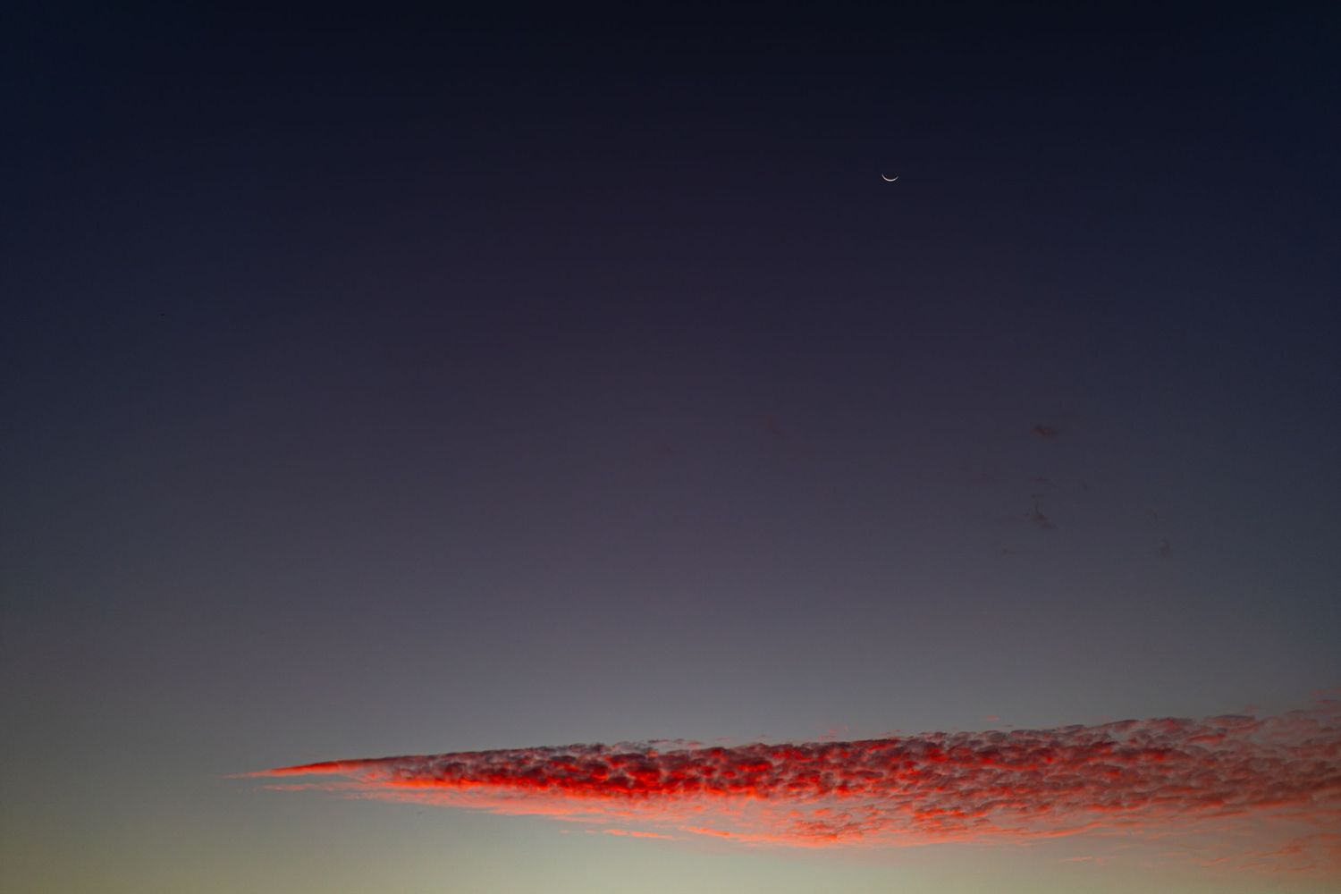Red-lit clouds streak across a twilight sky at dusk, with a crescent moon visible above the horizon.