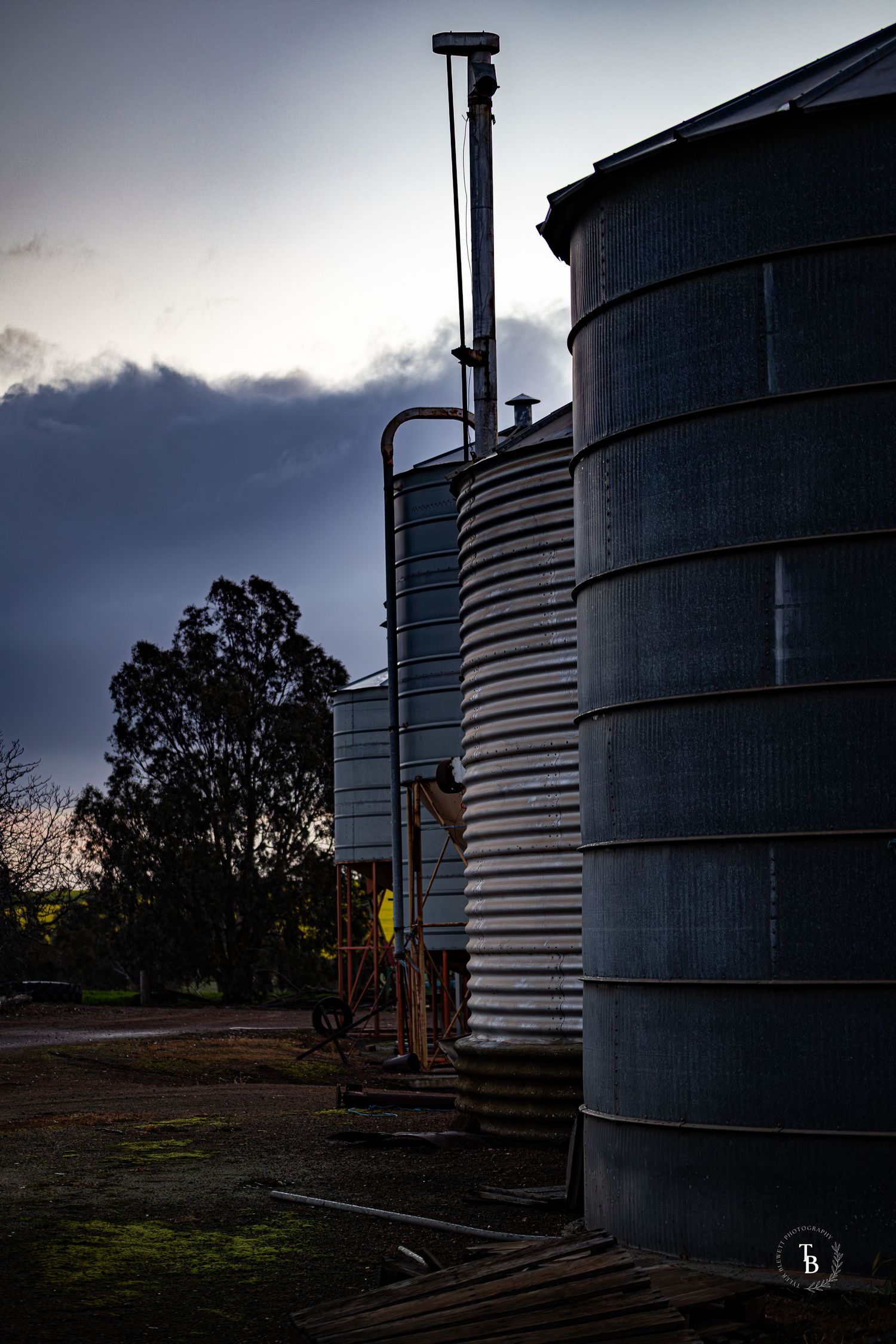 Tall corrugated metal grain silos stand against a moody dark cloudy sky at dusk on a farm.