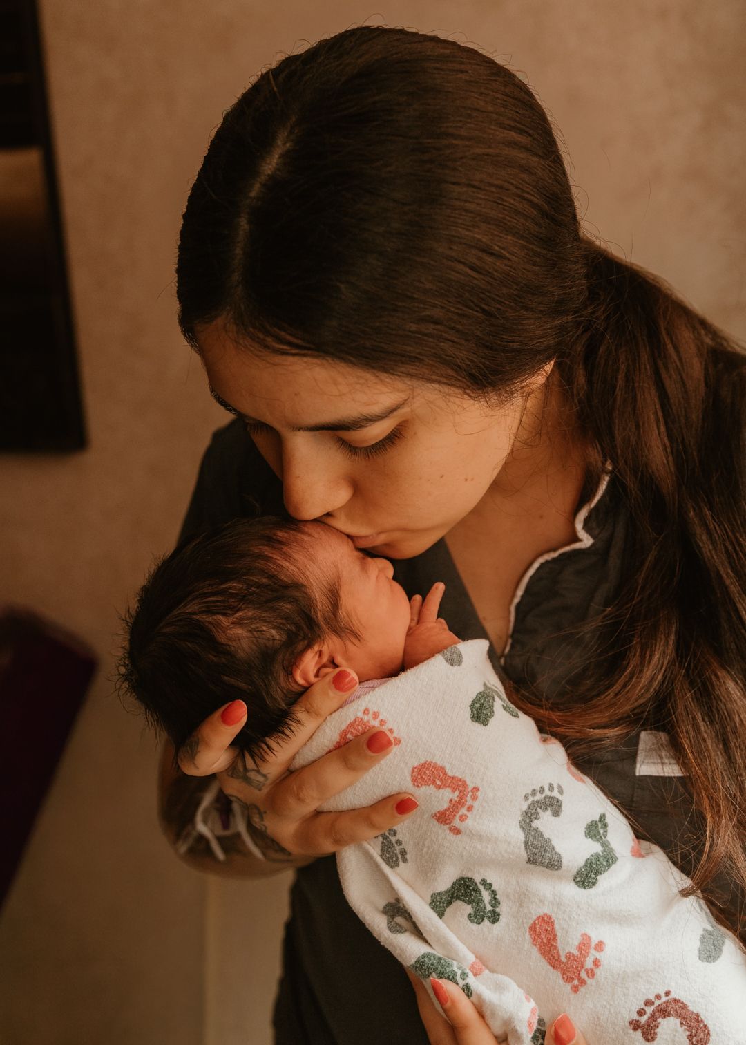 A mother tenderly kisses her newborn baby wrapped in a colorful footprint swaddle blanket.