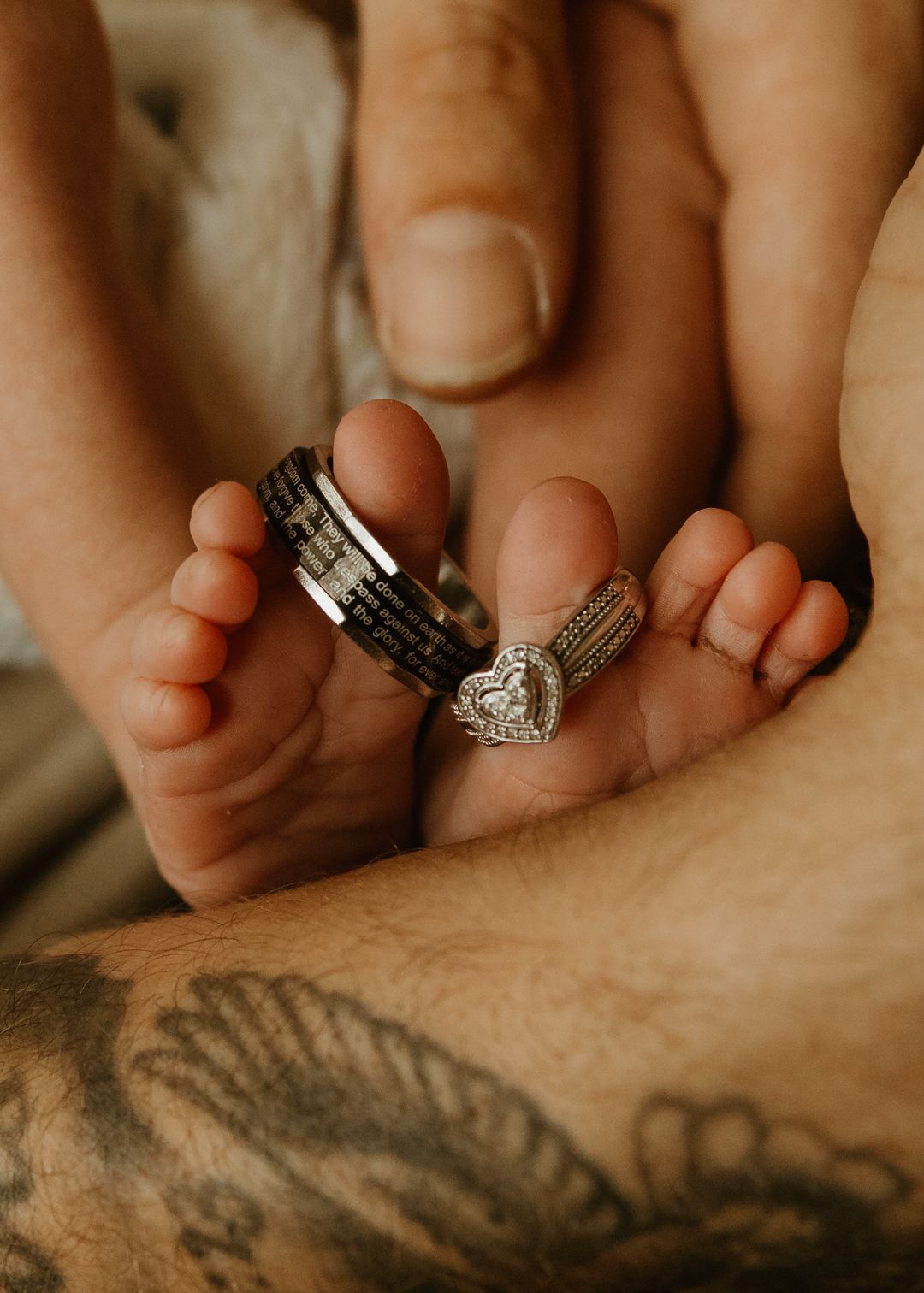 Baby feet adorned with wedding rings, held by parents hand with tattoo, newborn family photo.