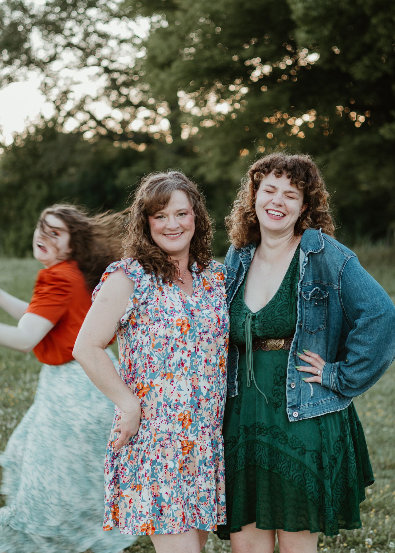Two curly-haired women smile outdoors in floral and green dresses, with a third woman in red visible behind them.