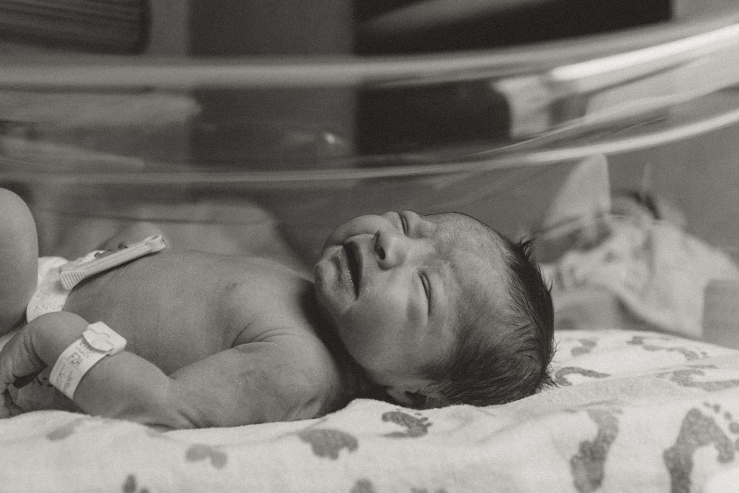 Black and white photo of a newborn baby resting in a hospital incubator with ID bracelet.