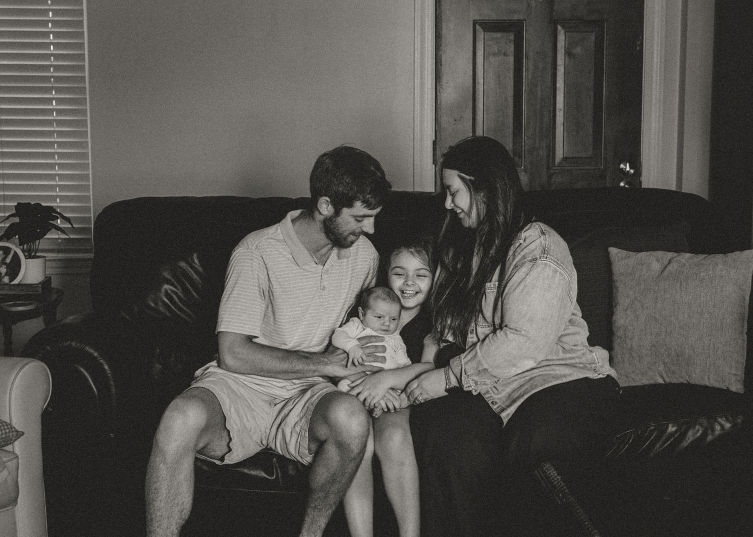 Black and white family portrait of parents and young child sitting together on a dark leather sofa at home.