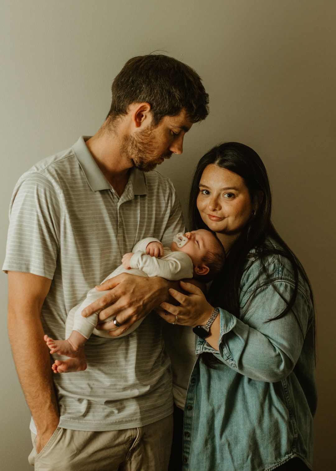 A young couple tenderly holds their newborn baby together, posing for a family portrait against a neutral background.