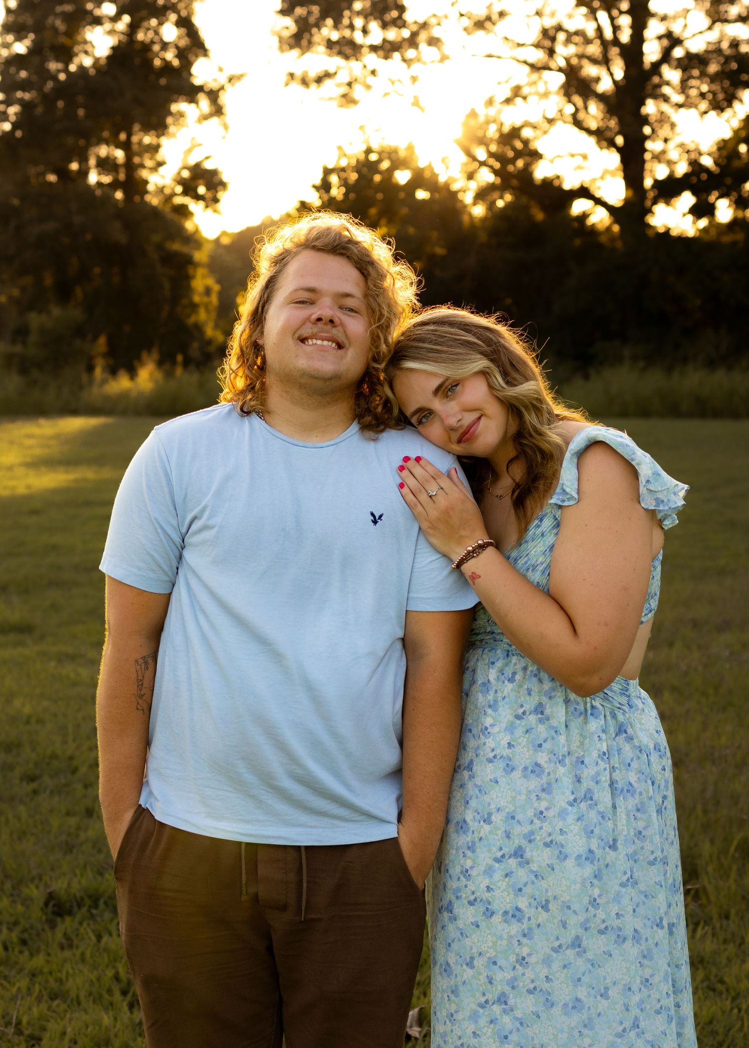 A young couple poses together at golden hour in a grassy field, woman in floral dress leaning on mans shoulder.