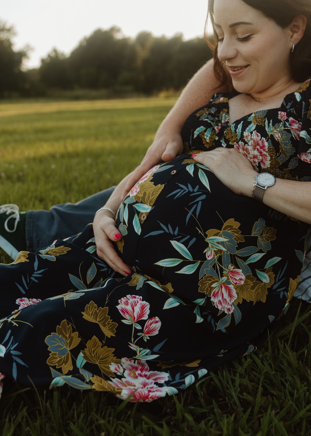 Pregnant woman in floral dress sitting in green field, cradling baby bump during golden hour maternity photo session.