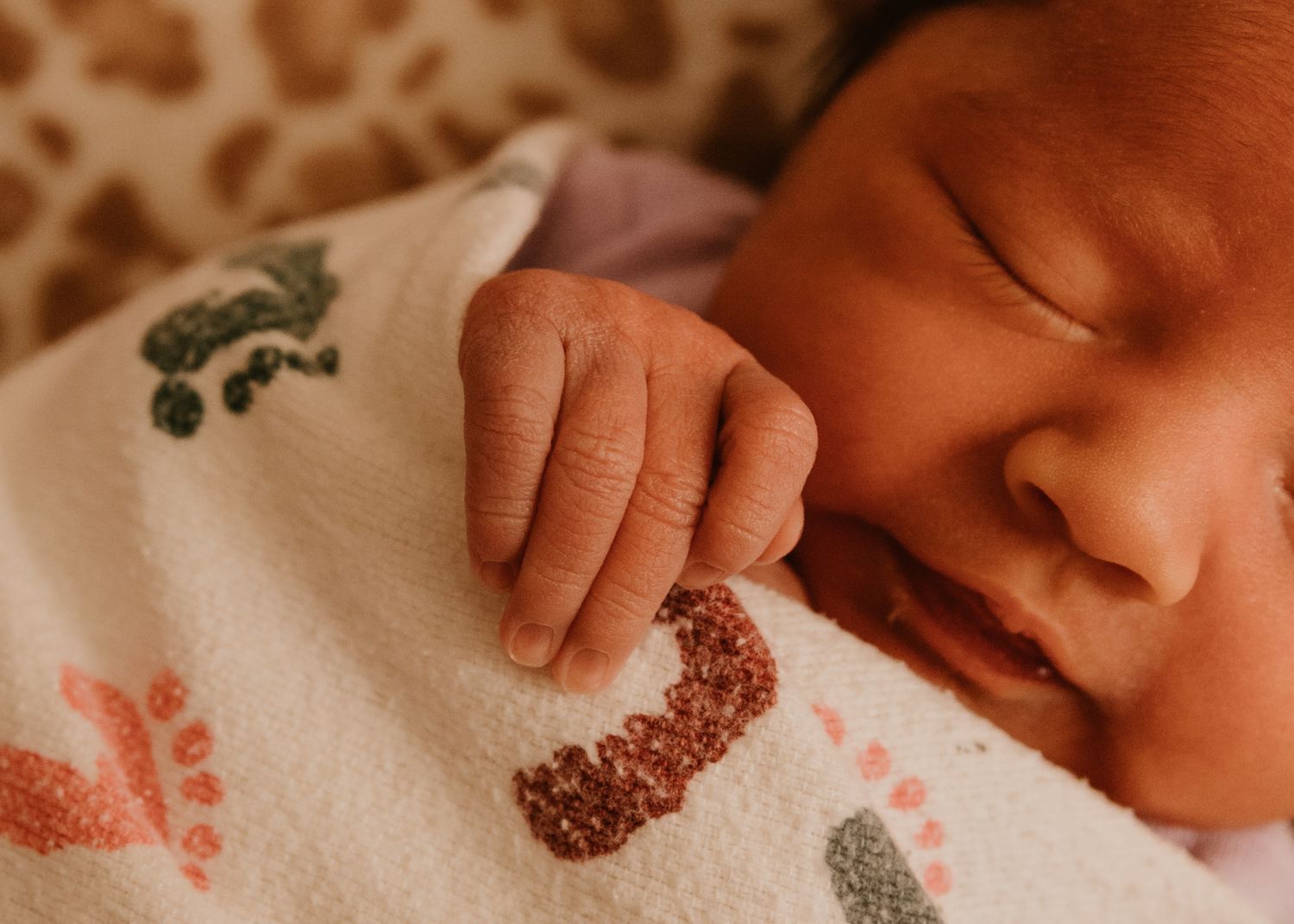Peaceful newborn baby sleeping swaddled in a white blanket with colorful prints, tiny hand visible.