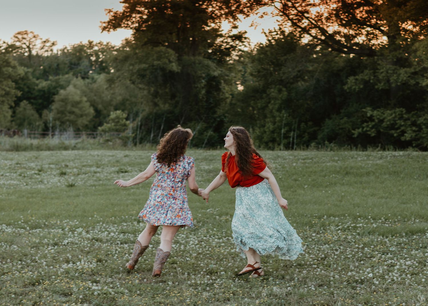 Two girls in floral dresses hold hands and dance together in a lush green meadow at golden hour.