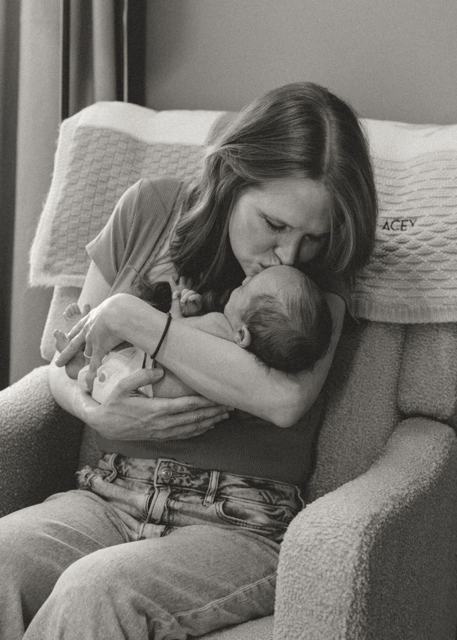 Black and white photo of a mother kissing her newborn baby while seated in a cozy armchair.