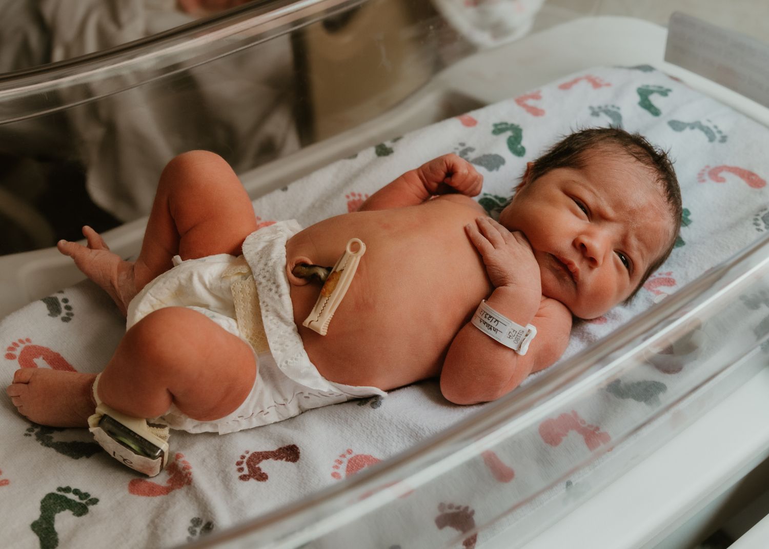 Newborn baby resting in hospital bassinet on colorful footprint blanket, wearing diaper and ID bracelet.