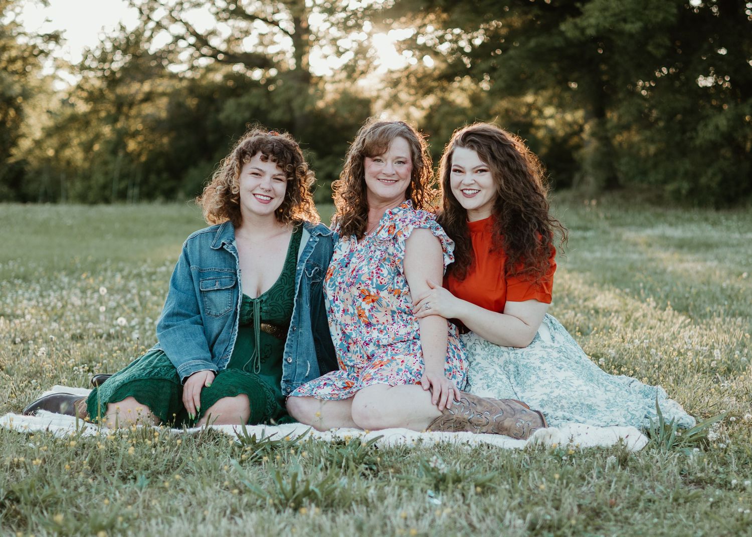 Three smiling young women sitting together on a blanket in a sunny green field during golden hour.