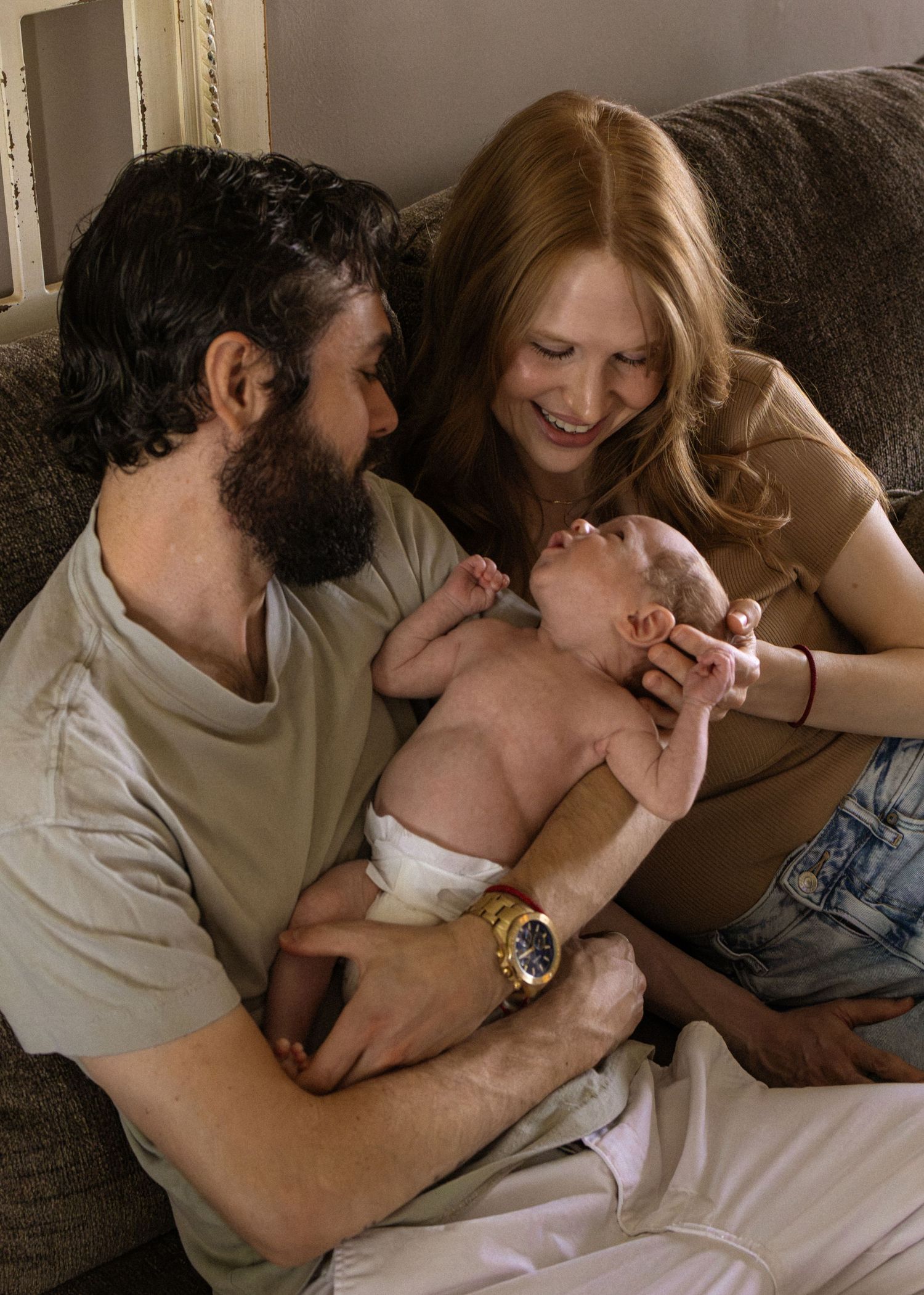 New parents sitting together on couch tenderly holding their newborn baby between them.