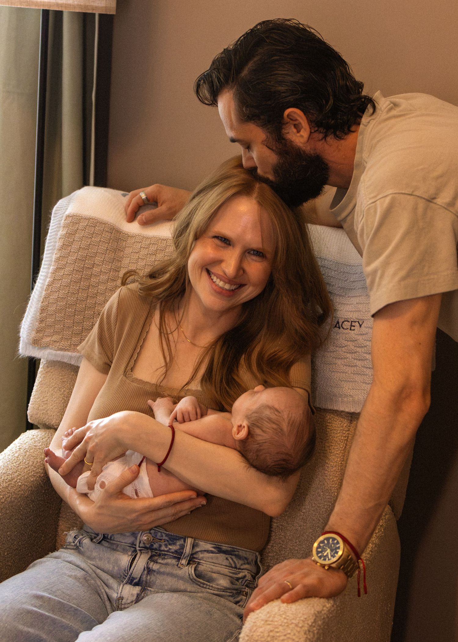 Happy mother holds newborn baby as bearded father leans in lovingly in a nursery chair.