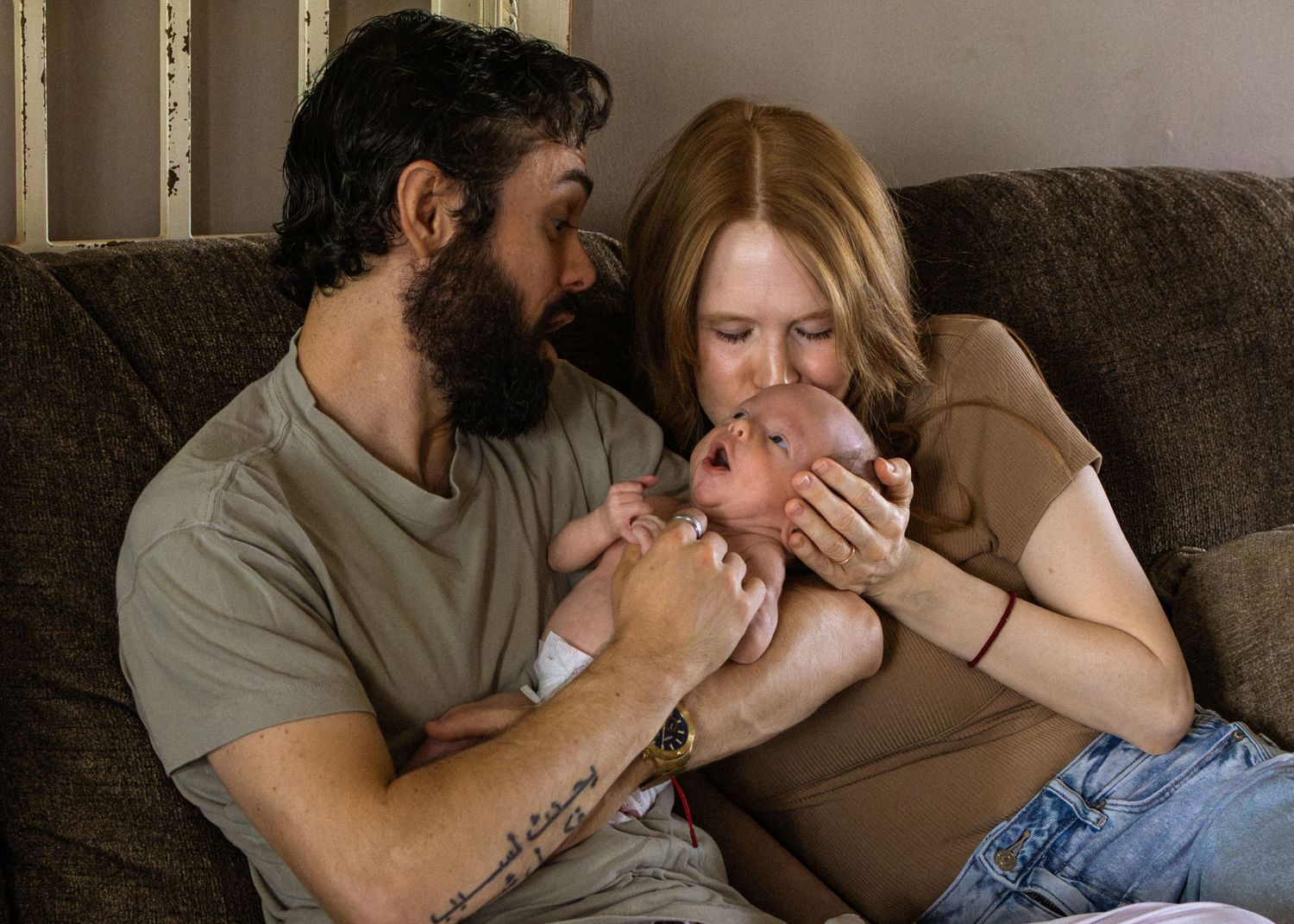 Bearded father and red-haired mother share a tender moment kissing their newborn baby.