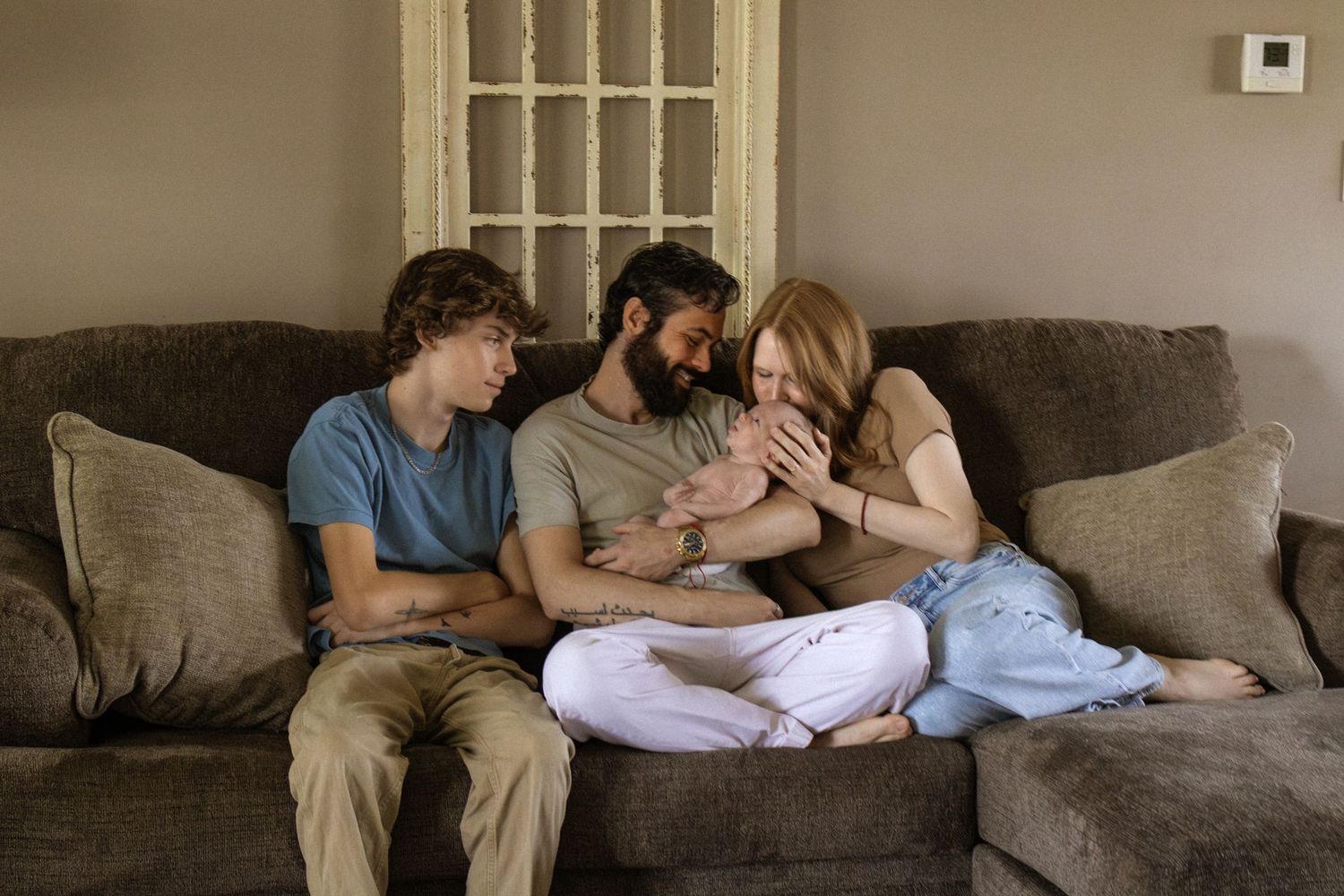 Family of three sits on brown couch admiring their newborn baby in a warm home setting.