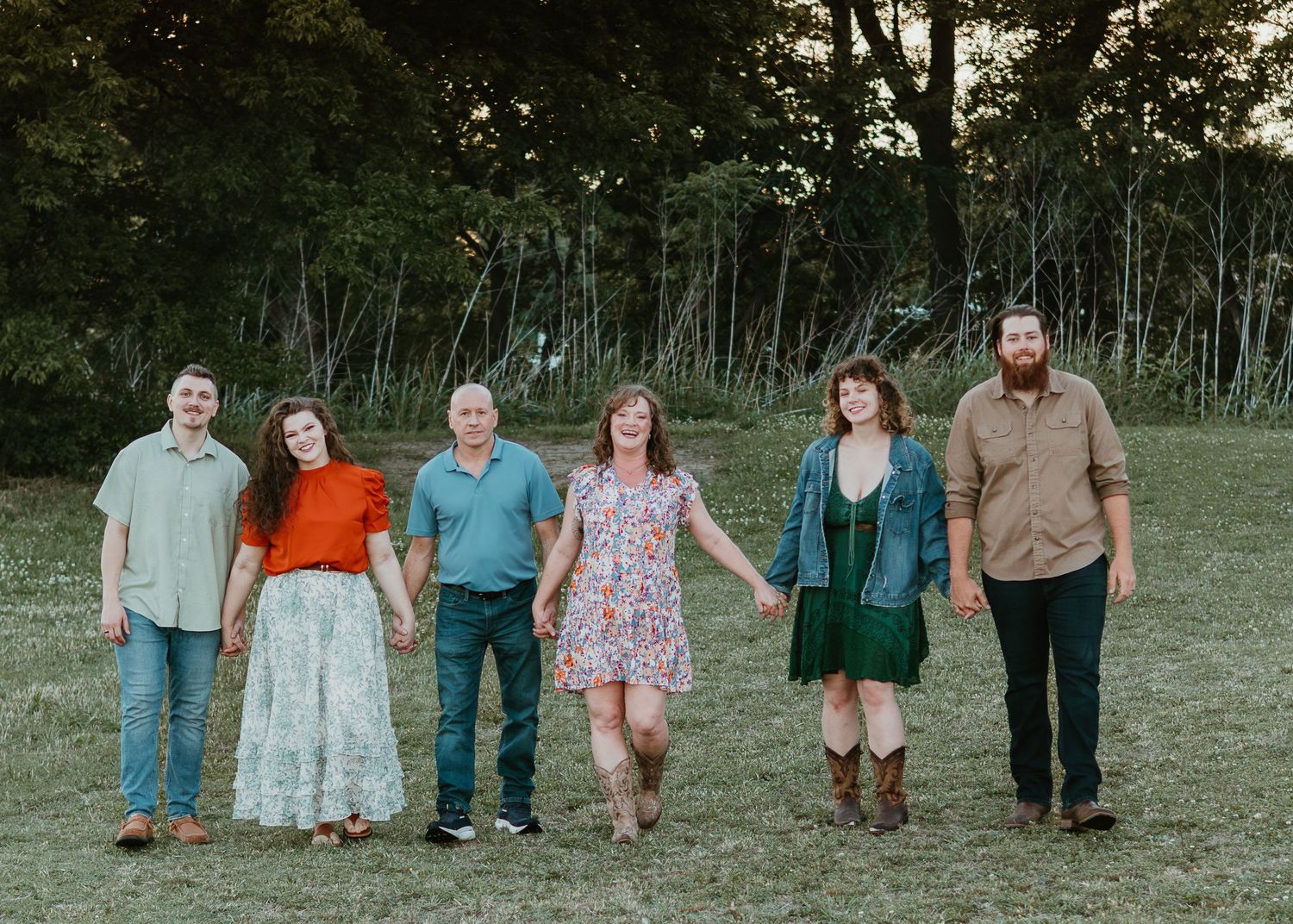 A family of six walks hand-in-hand across a green field with trees in the background during a portrait session.
