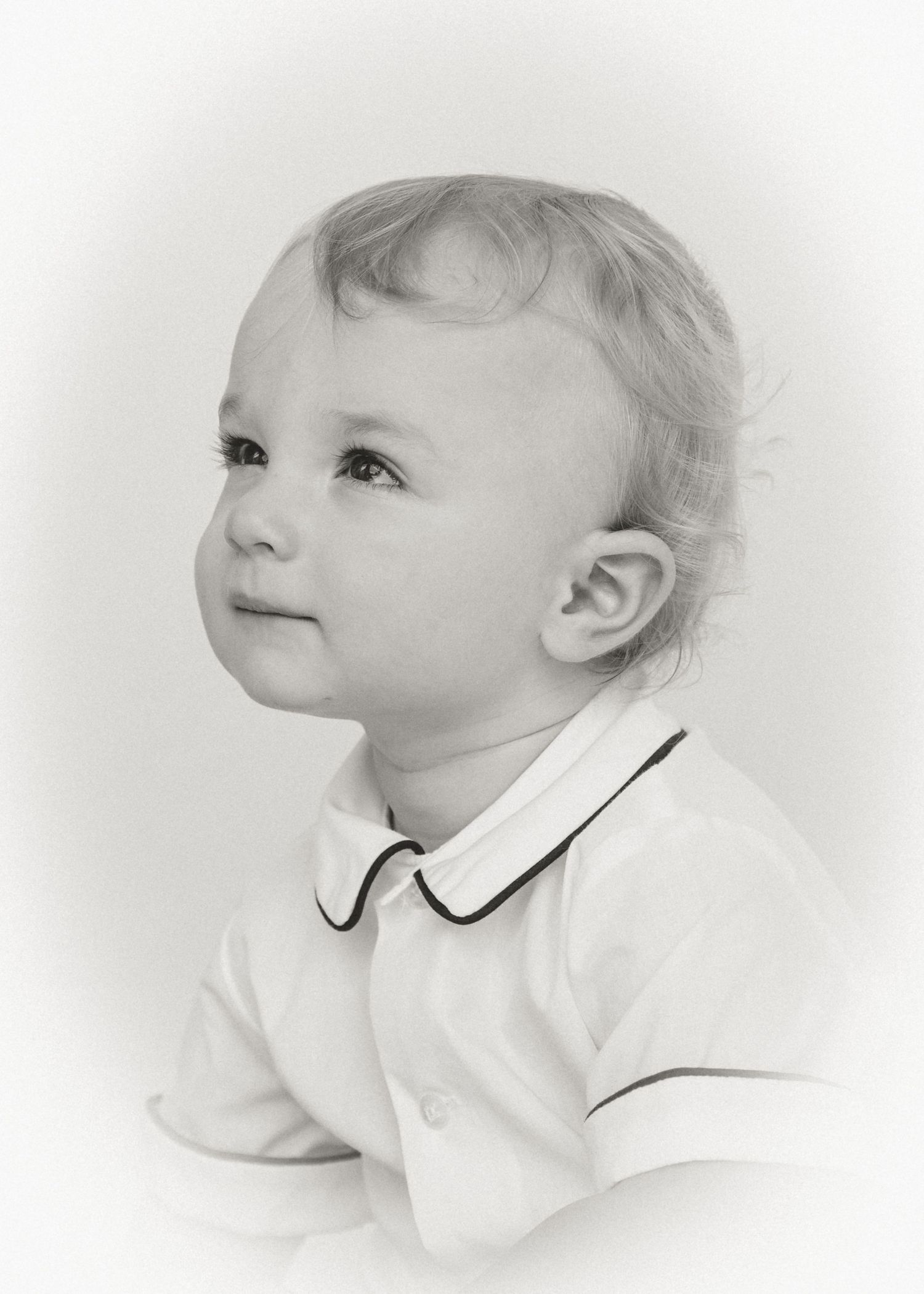 Black and white portrait of a cute toddler with light hair wearing a white polo shirt, looking upward.