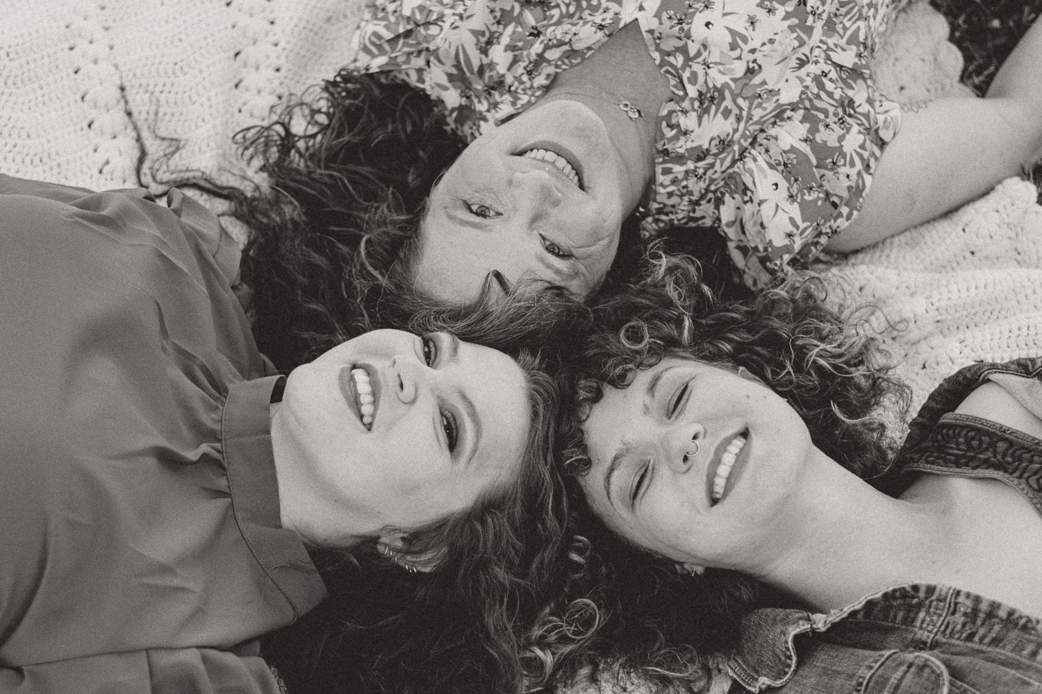 Three women with curly hair laughing together, lying down with heads touching in a black and white photo.