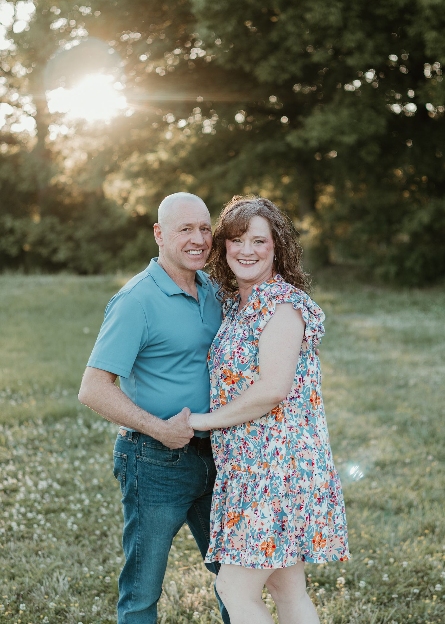 Couple smiling and embracing outdoors in a sunny field at golden hour, woman in floral dress, man in blue polo.