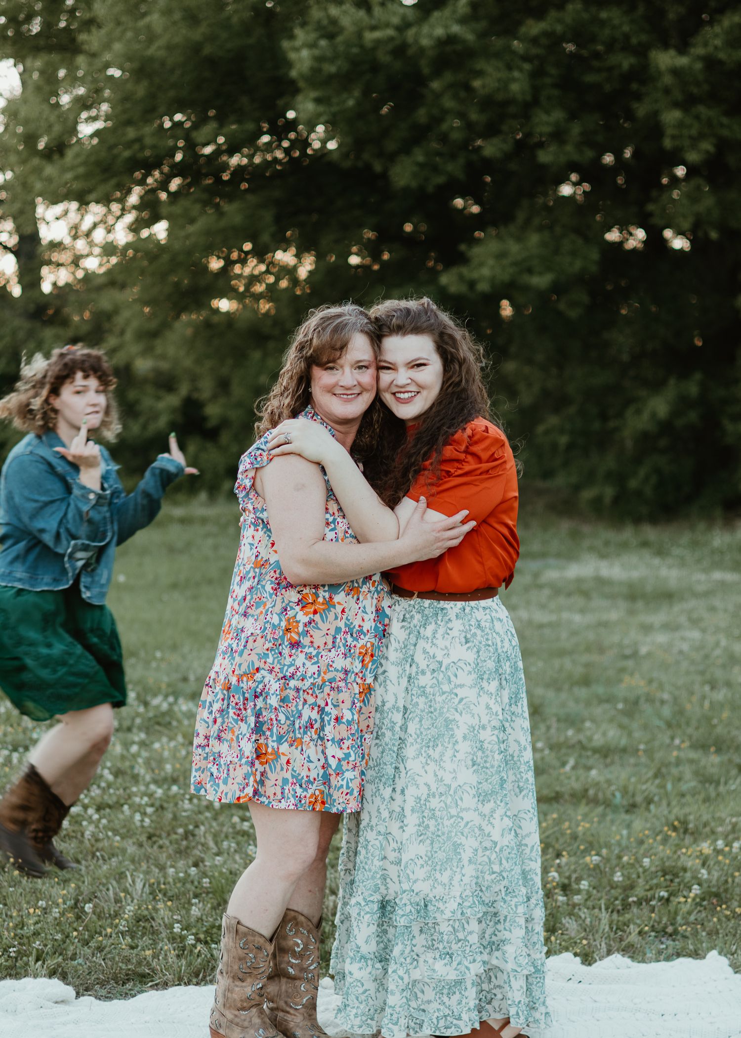 Two young women hugging and smiling in a green meadow, one in floral dress with cowboy boots, other in red jacket.