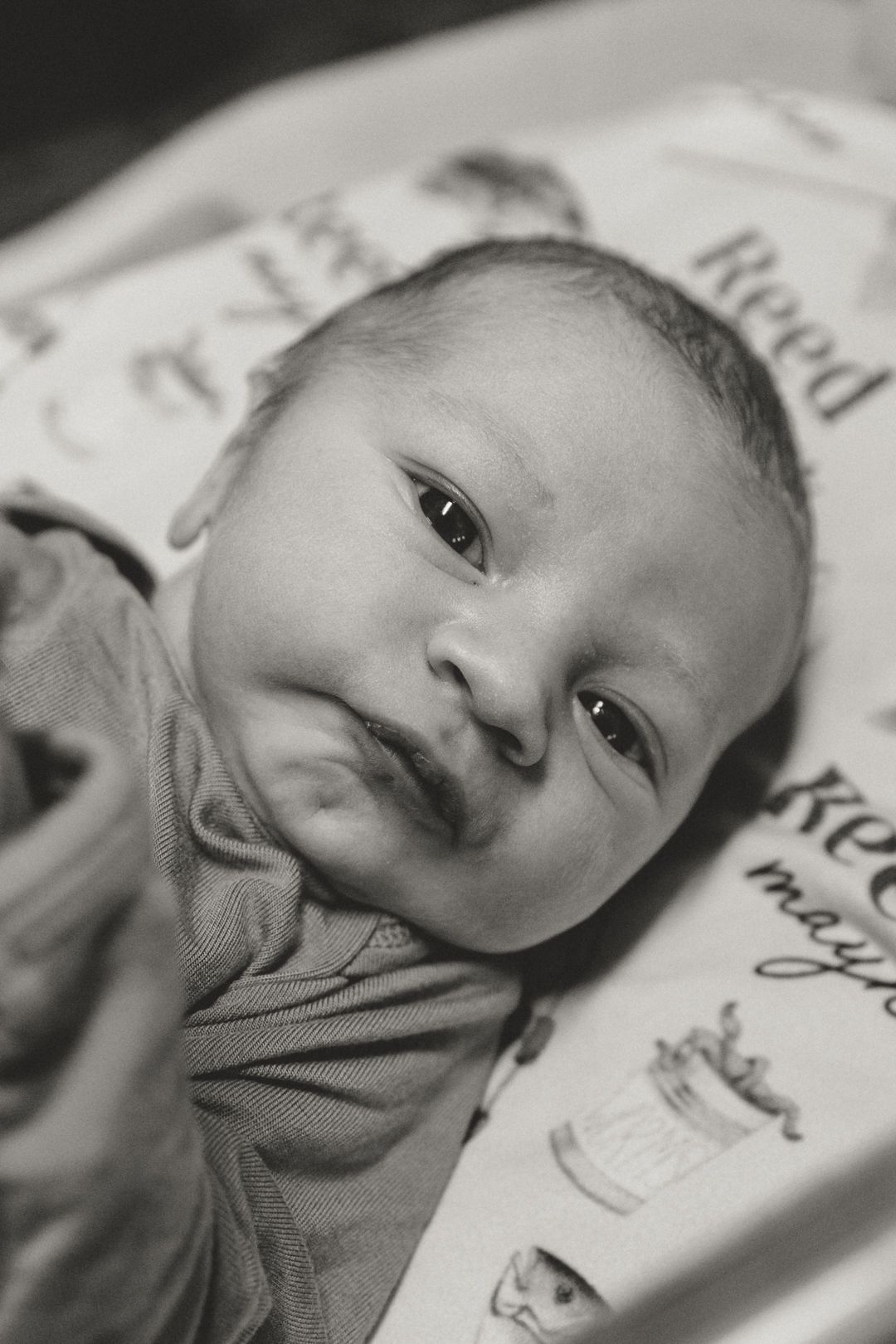 Black and white portrait of a newborn baby swaddled in a blanket, looking up with wide eyes and a subtle smile.