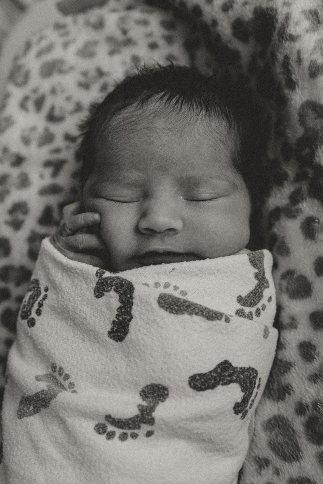 Black and white photo of a sleeping newborn baby swaddled in a white blanket with footprint patterns.