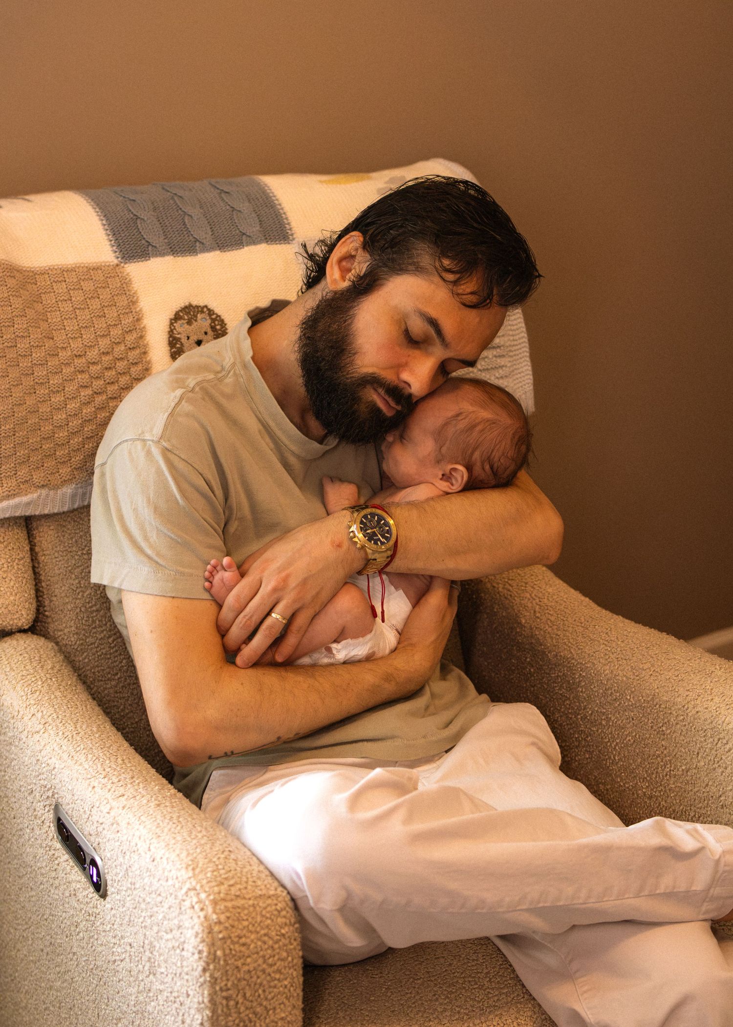 Bearded father sitting in a recliner chair gently cradling his sleeping newborn baby.