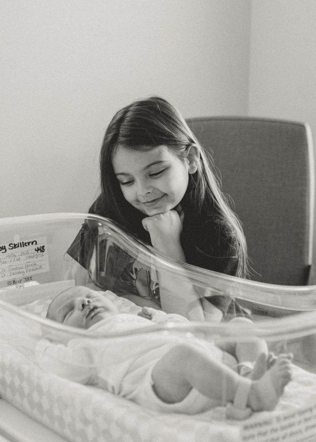 Black and white photo of a young girl smiling down at her newborn sibling in a hospital bassinet.