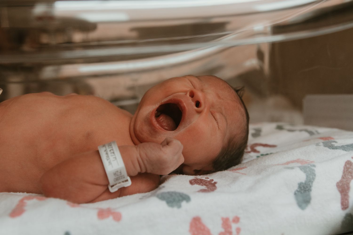 Newborn baby yawning in hospital nursery bassinet with ID bracelet on wrist, wrapped in colorful blanket.